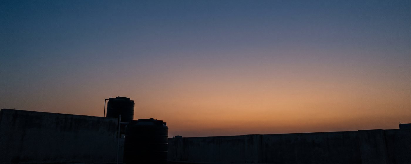 A thin crescent moon in the sky just before dawn, above a rooftop in silhouette