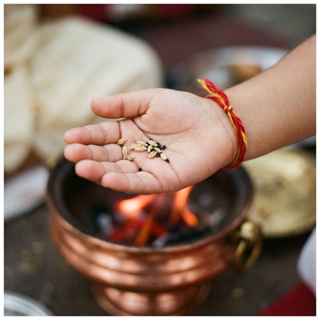 A child's palm holding barley and sesame above a small homa fire, sacred thread at the wrist