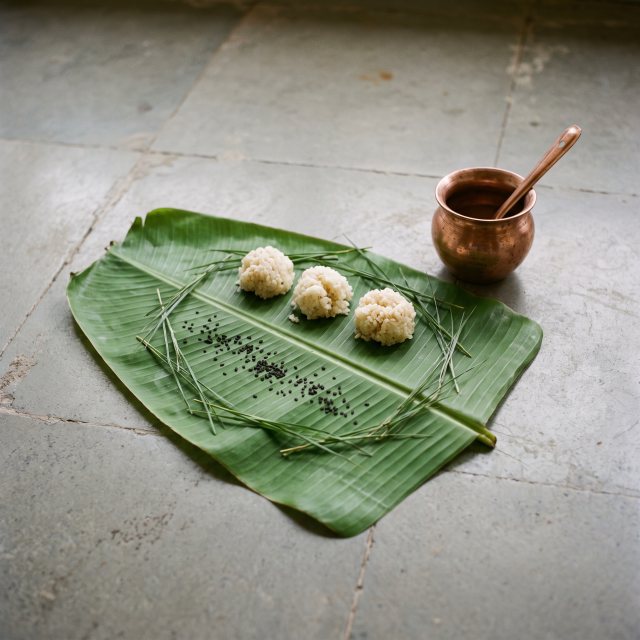 Rice offerings and black sesame on a banana leaf beside a copper water pot